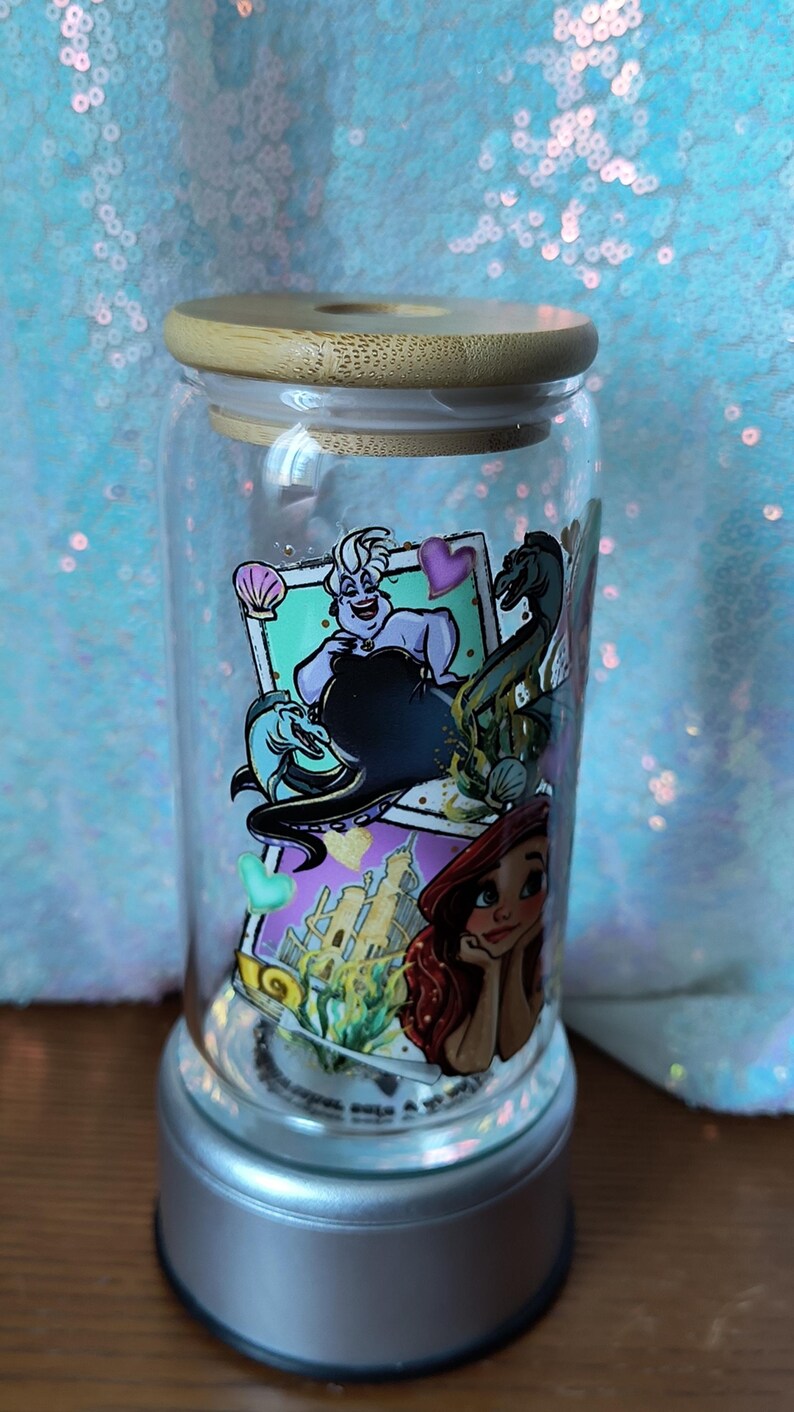 Op de afbeelding: Een doorzichtige glazen beker met een bamboe deksel, met kleurrijke illustraties van personages uit een populaire animatiefilm. De beker staat op een zilveren voet en is ongeveer 15 cm hoog.