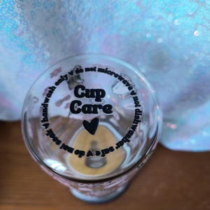 Op de afbeelding: Transparante glazen beker met de woorden "Cup Care" en een hartsymbool. De beker heeft tekst rond de rand met de tekst "handwash only", "do not soak", "do not microwave" en "dishwasher safe".