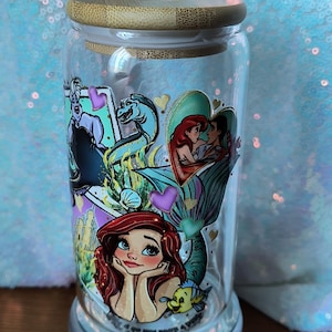 Op de afbeelding: Een heldere glazen pot met een bamboe deksel, met kleurrijke illustraties van personages uit "De Kleine Zeemeermin". De pot is versierd met afbeeldingen van Ariel, Ursula en andere personages. De pot staat op een zilveren voet.