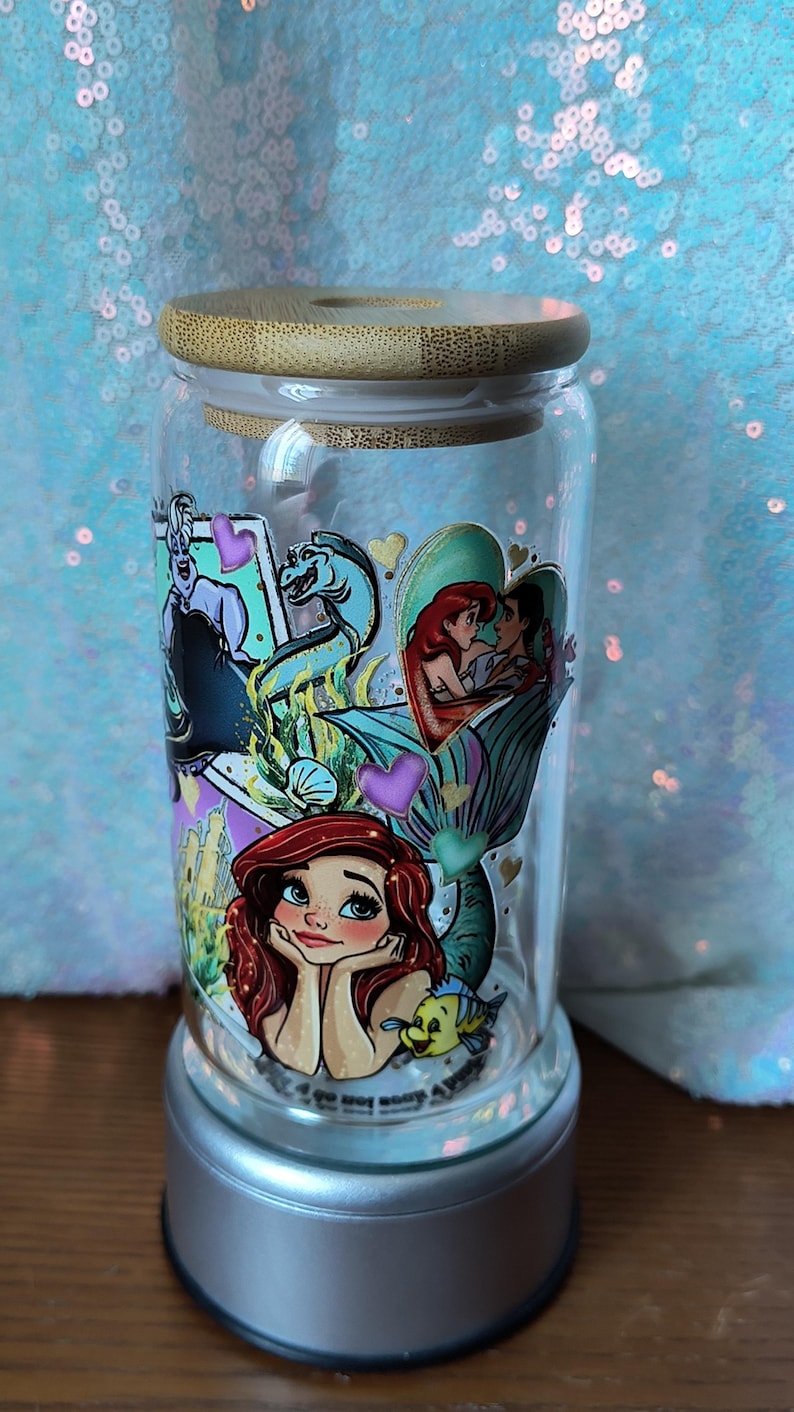 Op de afbeelding: Een heldere glazen pot met een bamboe deksel, met kleurrijke illustraties van personages uit "De Kleine Zeemeermin". De pot is versierd met afbeeldingen van Ariel, Ursula en andere personages. De pot staat op een zilveren voet.