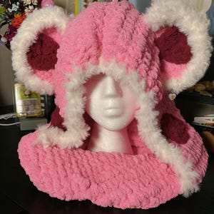 May include: A pink, plush, handmade hood with large, rounded ear-like features. The hood is trimmed with white faux fur and has burgundy accents inside the ears. The hood is designed to be worn around the head and neck.