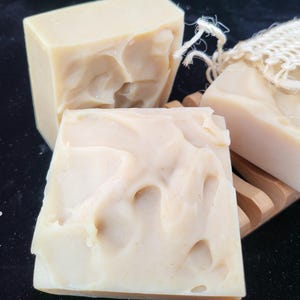 May include: Three bars of handmade soap, two are white and one is a light beige color. The soap bars are on a wooden surface.