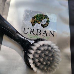 May include: A black facial brush with white bristles. The brush is next to a silver label with the words "URBAN Goddess 330" and a graphic of a woman's face with green and gold hair.