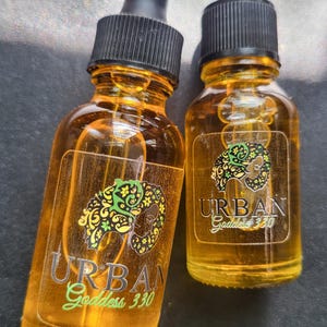 May include: Two amber glass bottles with dropper tops, both labeled "URBAN Goddess 330". The labels feature a stylized image of a woman's face with floral designs.