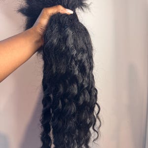 Peut inclure: Une mèche d'extensions de cheveux noirs et ondulés. Les cheveux sont maintenus ensemble en haut, avec les mèches ondulées qui descendent. La texture semble être grossière et naturelle, adaptée à diverses coiffures.