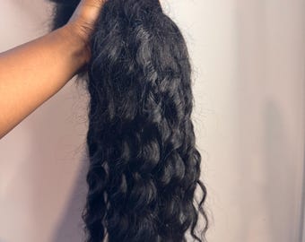 May include: A bundle of black, wavy hair extensions. The hair is held together at the top, with the wavy strands cascading downwards. The texture appears to be coarse and natural-looking, suitable for various hairstyles.