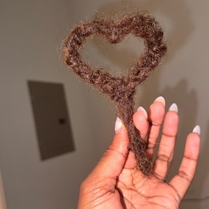 May include: A heart shape made of brown dreadlocks. The heart is held up by a hand with manicured nails. The background is a neutral wall with a rectangular panel.