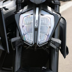 Puede incluir: Primer plano de una motocicleta KTM negra. La parte delantera de la motocicleta presenta un diseño de faro distintivo con el logotipo de KTM. Se ven el manillar y la suspensión delantera, mostrando el diseño elegante y moderno de la moto.