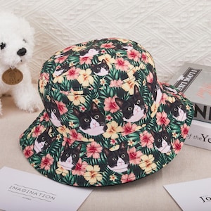 May include: A patterned bucket hat featuring black and white cat faces, pink and yellow flowers, and green foliage. The hat is made of a lightweight material and has a wide brim.
