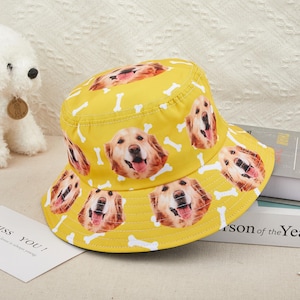 May include: A bright yellow bucket hat adorned with a repeating pattern of golden retriever faces and white bone motifs. The hat, with its wide brim, is designed for sun protection. A cheerful accessory for dog enthusiasts, ideal for sunny days.