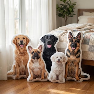 May include: Five dog-shaped throw pillows featuring different breeds: a Golden Retriever, French Bulldog, Black Labrador, Maltese, and German Shepherd. They are arranged on a wooden floor near a window and a bed with a patterned quilt.