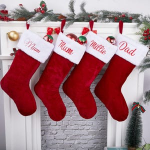 May include: Four red Christmas stockings with white faux fur cuffs hang from a mantelpiece. Each stocking is embroidered with a name: "Mum", "Mum", "Millie", and "Dad". The stockings are decorated with small baubles and bows, with a small Christmas tree in the background.