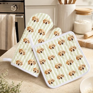 May include: A quilted oven mitt and a square pot holder featuring a repeating pattern of golden retriever dog faces on a light sage green and white diagonal striped background with white fabric trim along the edges of the kitchen accessories.