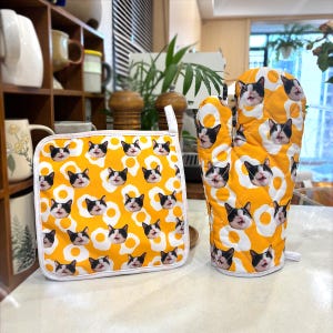 May include: A yellow oven glove and pot holder set. The set features a repeating pattern of cat faces and fried eggs. The cat faces are black and white, and the eggs are white with yellow yolks. The pot holder has a white border and a hanging loop.