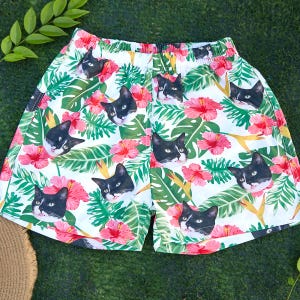Puede incluir: Pantalones cortos blancos con un estampado tropical vibrante con caras de gatos en blanco y negro, flores de hibisco rosa y hojas verdes. Los pantalones cortos tienen una cintura elástica y están hechos de tela ligera.