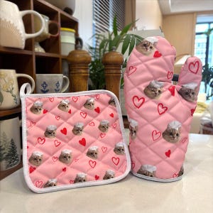 May include: Pink quilted oven mitt and pot holder set. The set features a repeating pattern of chef cat faces and red hearts. The oven mitt has a thumb and four finger slots. The pot holder is square with a white border.
