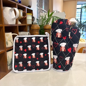 Puede incluir: Un juego de manopla y agarradera negros. El juego presenta un patrón repetido de una persona sonriente con un gorro de chef y corazones rojos. Los artículos tienen un ribete blanco y están sobre una superficie clara.