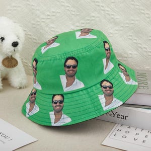 May include: A green bucket hat featuring a repeating pattern of a person wearing sunglasses and a white shirt. The hat has a black brim and is sitting on a white surface. The hat is a casual accessory.