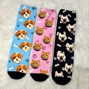 May include: Three pairs of colourful socks with different pet faces printed on them. The socks are blue, pink, and black with white paw prints and bone designs.