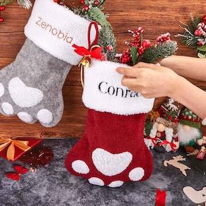 Peut inclure: Deux bas de Noël en forme d'empreintes de pattes. L'un est gris avec des accents blancs et le nom "Zenobia" brodé sur le revers. L'autre est rouge avec des accents blancs et le nom "Conrad" brodé sur le revers. Les deux ont un revers blanc duveteux.