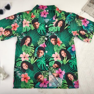 Puede incluir: Camisa de manga corta con estampado tropical de hojas verdes, flores rosas y caras sobre fondo negro. La camisa tiene cuello de camisa y botones blancos. Estilo abotonado.
