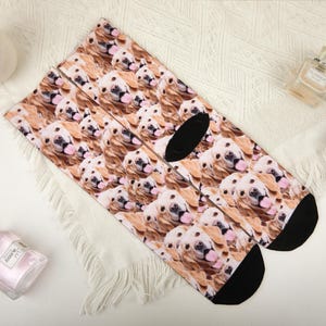 May include: A pair of socks with a repeating pattern of golden retriever faces. The socks have black heels and toes. The background is white.