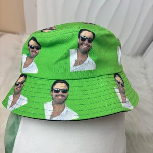 May include: Green bucket hat with a black brim and a repeated pattern of a man wearing sunglasses.