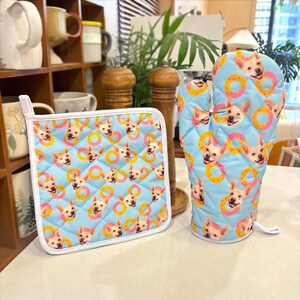 May include: A light blue quilted oven mitt and pot holder set. The set features a repeating pattern of a dog's face and yellow and pink inflatable rings. The pot holder has a white border and a hanging loop.