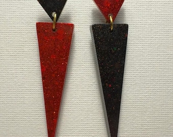 Könnte beinhalten: Ein Paar lange, dreieckige Ohrringe mit schwarzem und rotem Glitzerfinish. Die Ohrringe bestehen aus zwei Teilen, einem kleineren Dreieck oben und einem größeren Dreieck unten. Das obere Dreieck ist schwarz und das untere Dreieck ist rot.