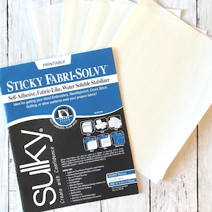 May include: A package of Sulky Sticky Fabri-Solvy, a self-adhesive, fabric-like, water-soluble stabilizer for hand embroidery, needlepunch, cross stitch, and quilting. The package is blue and white, with a stack of white sheets next to it. The sheets measure 12 x 11 inches.