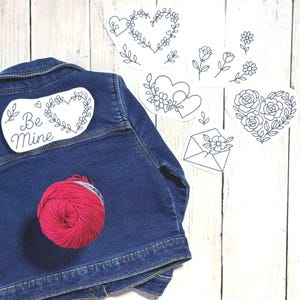 May include: A dark blue denim jacket with a red ball of yarn and several white paper cutouts featuring black line drawings of hearts, flowers, and an envelope. One cutout says "Be Mine".