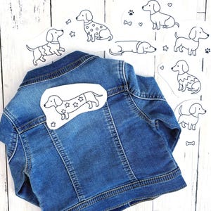 Puede incluir: Una chaqueta de mezclilla azul con un parche blanco de un perro salchicha con patrones de estrellas en el lomo. Alrededor de la chaqueta hay varias pegatinas blancas con ilustraciones de líneas negras de perros salchicha en varias poses.