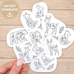 May include: A collection of white paper cutouts featuring black line art illustrations of woodland animals including a bear, deer, foxes, rabbits, a squirrel, and birds, each decorated with floral patterns and leafy sprigs.