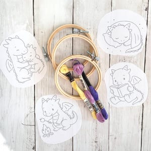 May include: Embroidery kit featuring dragon designs. Includes wooden embroidery hoops, embroidery floss in yellow and purple, and printed fabric with dragon illustrations. The designs include a dragon reading, sleeping, hugging, and roasting marshmallows.