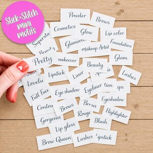 May include: A collection of small white rectangular paper labels featuring black cursive text related to makeup and beauty, including words like Powder, Brows, Cosmetics, Glow, Lip color, Foundation, Glam, Beauty, Lipstick, Liner, Lashes, and Blush.