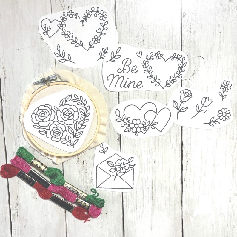 May include: Embroidery kit with heart and floral designs. Includes a wooden embroidery hoop with a heart-shaped pattern, pre-printed designs, and spools of red, pink, and green embroidery floss. The designs include the text "Be Mine".