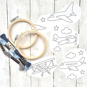 Puede incluir: Kit de bordado con aros de madera, hilo de bordar azul y diseños de aviones. Incluye cuatro contornos de aviones, nubes y estrellas. El kit está sobre un fondo de vetas de madera clara.