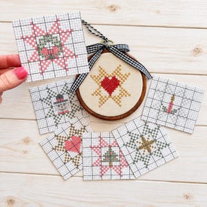 May include: A circular wooden embroidery hoop containing a cross-stitch heart pattern, accented with a black and white gingham ribbon. Surrounding the hoop are six square paper pattern charts featuring cross-stitch designs of stars, a tree, and a candle.