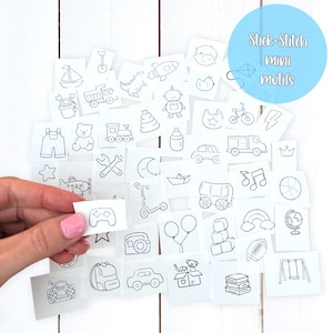 May include: A collection of white, rectangular Stick+Stitch mini motifs with black line drawings of various objects, including a boat, a rocket, a car, and a teddy bear. A hand holds one motif with a game controller design.