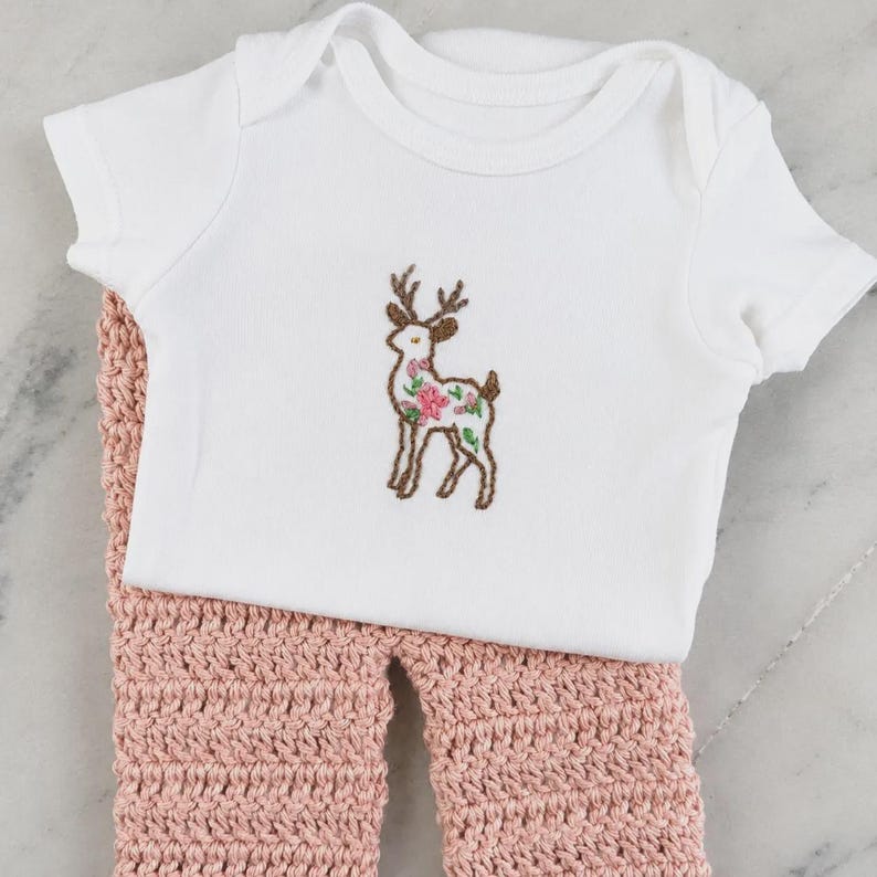 May include: A white short-sleeved bodysuit featuring a brown embroidered deer outline filled with pink and green floral patterns, paired with dusty pink crocheted pants that have a textured, thick yarn stitch throughout the garment.