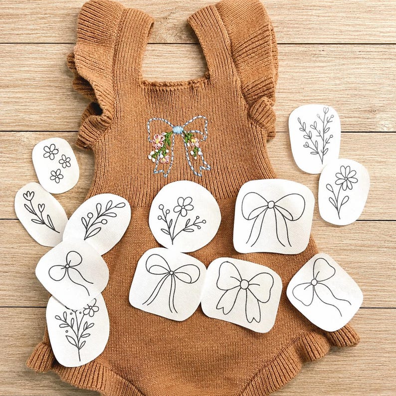 May include: A tan knit romper with ruffled sleeves and a central embroidered bow featuring small floral details. The garment is surrounded by several white paper cutouts displaying black line drawings of various bows and floral sprigs.