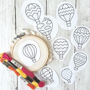 May include: An embroidery hoop with a hot air balloon design, surrounded by several other hot air balloon templates. Colorful embroidery floss in orange, yellow, and pink is also present, suggesting a crafting project. The designs are outlined in black.