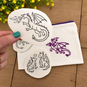 May include: A white canvas zipper pouch featuring a purple embroidered dragon, accompanied by two circular white fabric patches. One patch displays a black embroidered dragon breathing fire, while the other shows two small dragons facing each other.