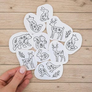May include: A collection of white stickers featuring black line art illustrations of various forest animals including a bear, deer, fox, squirrel, rabbit, and birds, each decorated with delicate floral and botanical patterns on their bodies.