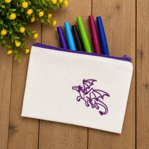 May include: A white canvas zippered pouch featuring a purple embroidered dragon design. The pouch has a purple zipper closure and contains five colorful pens in shades of purple, blue, black, green, and magenta, partially visible inside the top.