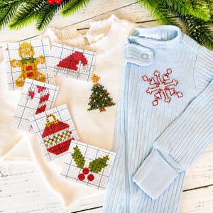 May include: A white sweater featuring a small embroidered Christmas tree, a light blue ribbed onesie with a red snowflake embroidery, and five grid-patterned cards displaying cross-stitch designs of a gingerbread man, hat, candy cane, ornament, and holly.