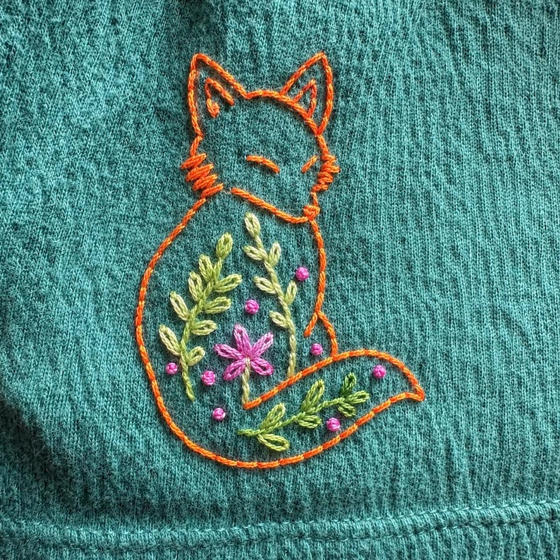 May include: A teal fabric garment featuring an orange embroidered outline of a fox. The fox body is filled with green embroidered leafy stems, a central pink flower, and small pink French knot accents scattered throughout the design.