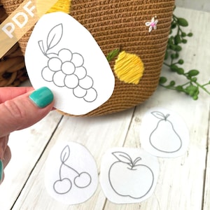 May include: A hand holding a white paper cutout of a bunch of grapes, with a woven basket in the background. Other fruit cutouts include a pear, cherries, and an apple. The basket has yellow embroidery and a small white flower.
