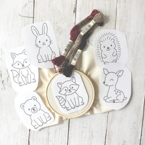 May include: Embroidery kit featuring woodland animal designs. Includes a wooden hoop, fabric, embroidery floss in shades of red, brown, and black, and pre-printed patterns of a fox, bear, rabbit, raccoon, deer, and hedgehog.