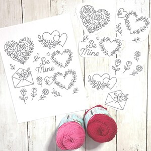 May include: Valentine's Day-themed line art illustrations featuring hearts, roses, and the words "Be Mine." Two balls of yarn, one pink and one red, are also visible. The illustrations are on white paper, set against a light wooden background.